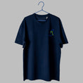Load image into Gallery viewer, A navy blue t-shirt on a blue wire hanger against a light grey background. It features a small embroidered design on the left chest showing a blue outline of a foot wearing a shoe with bright green laces.