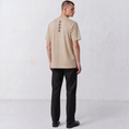 Charger l'image dans la visionneuse de la galerie, Three-quarter back view of a model wearing the beige Gordina Unsigned T-shirt with burgundy cross-stitch embroidery.