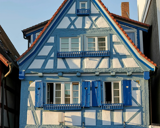 Charming half-timbered house with blue and white painted wooden beams and shutters, featuring a steep red-tiled roof and traditional European architecture, captured in warm sunlight.