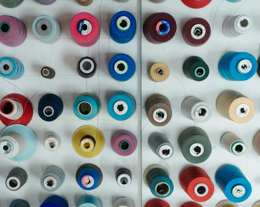 Top-down view of multiple colorful spools of thread neatly arranged on a white wall-mounted rack, showcasing a variety of textures and shades including red, blue, grey, yellow, and green.