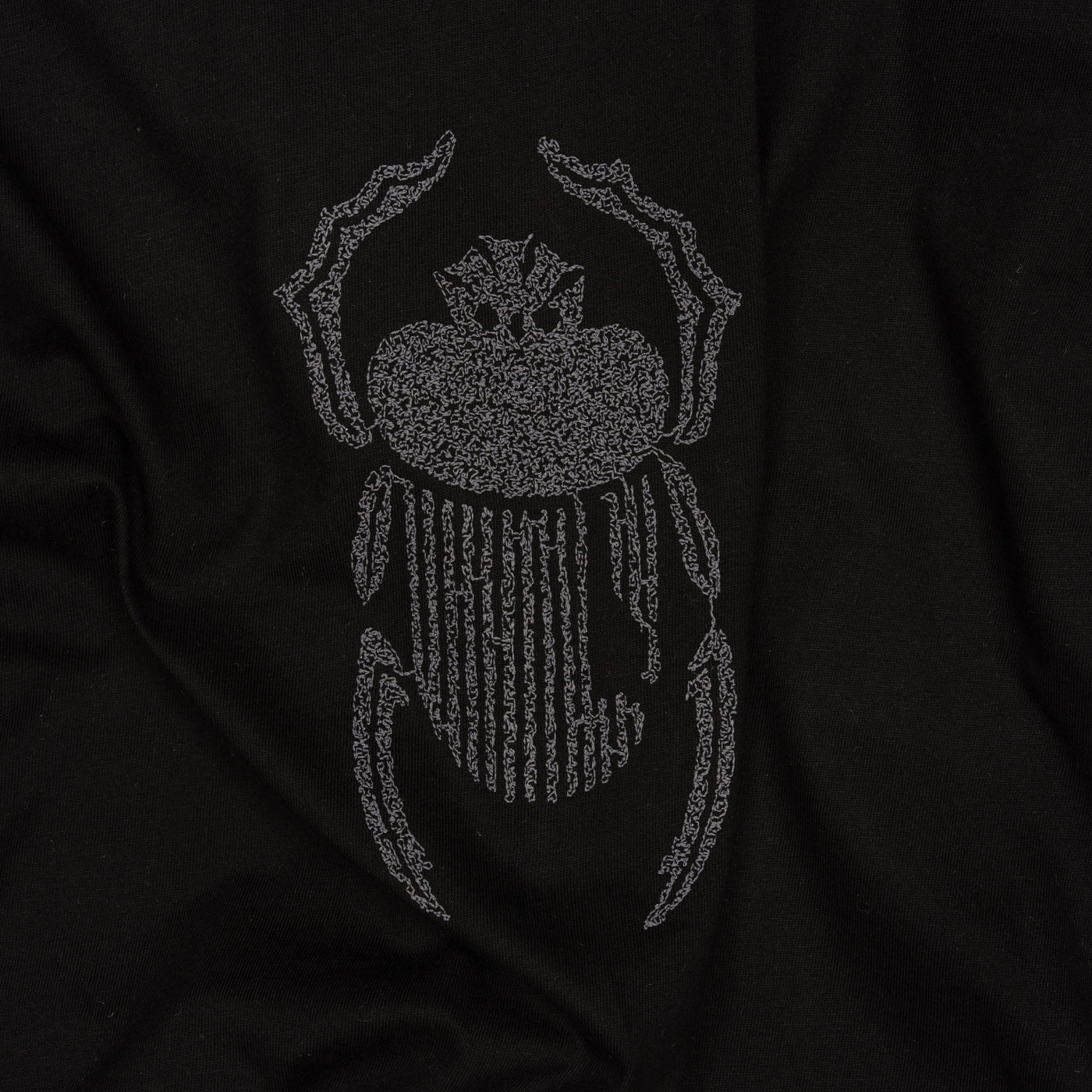 Wide close-up of grey scarab embroidery stitched onto the Gordina black oversized cotton T-shirt.