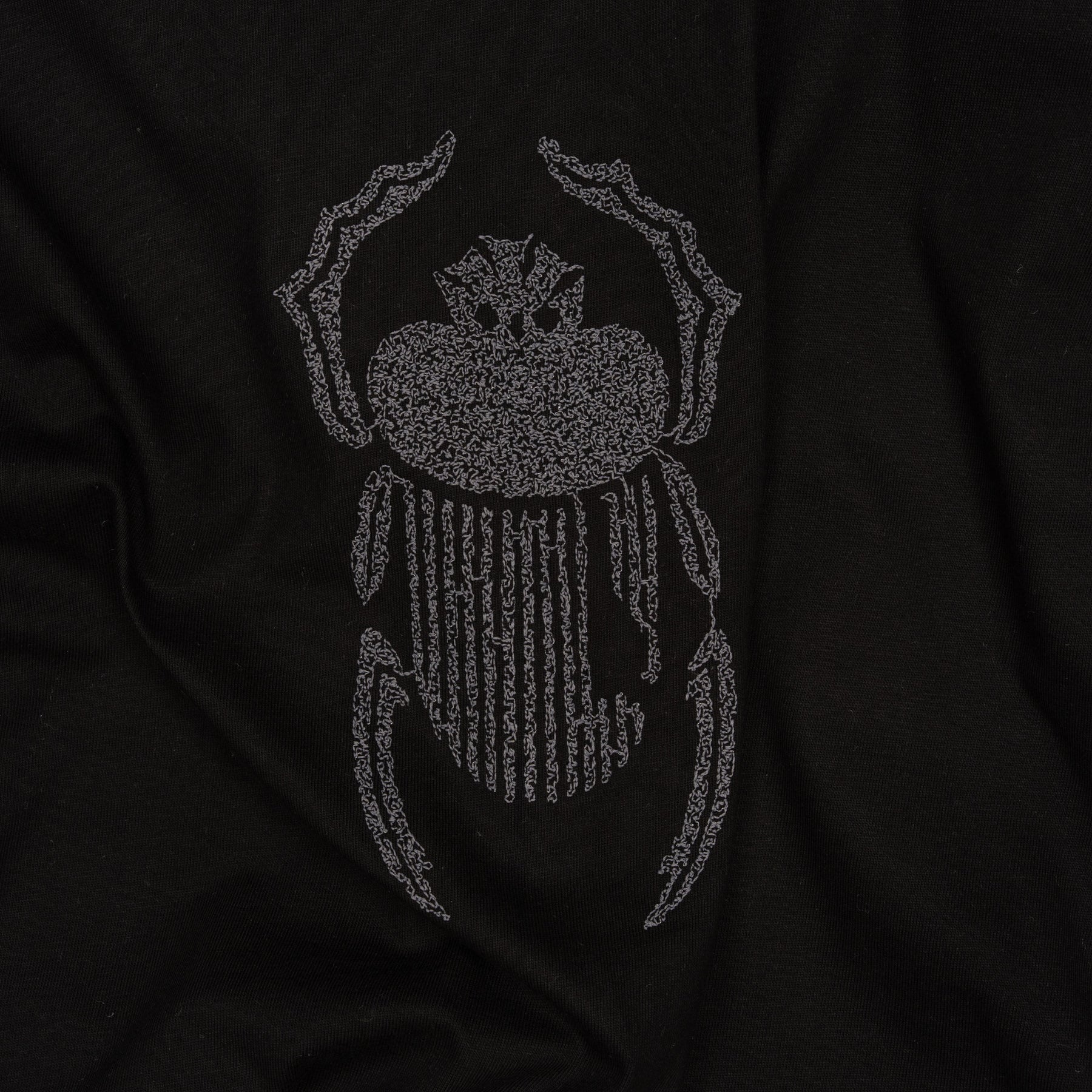 Wide close-up of grey scarab embroidery stitched onto the Gordina black oversized cotton T-shirt.