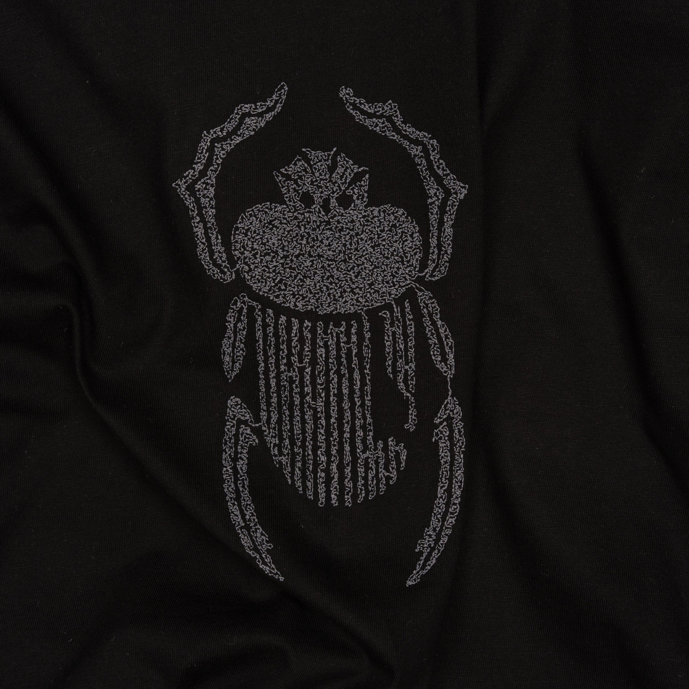 Wide close-up of grey scarab embroidery stitched onto the Gordina black oversized cotton T-shirt.