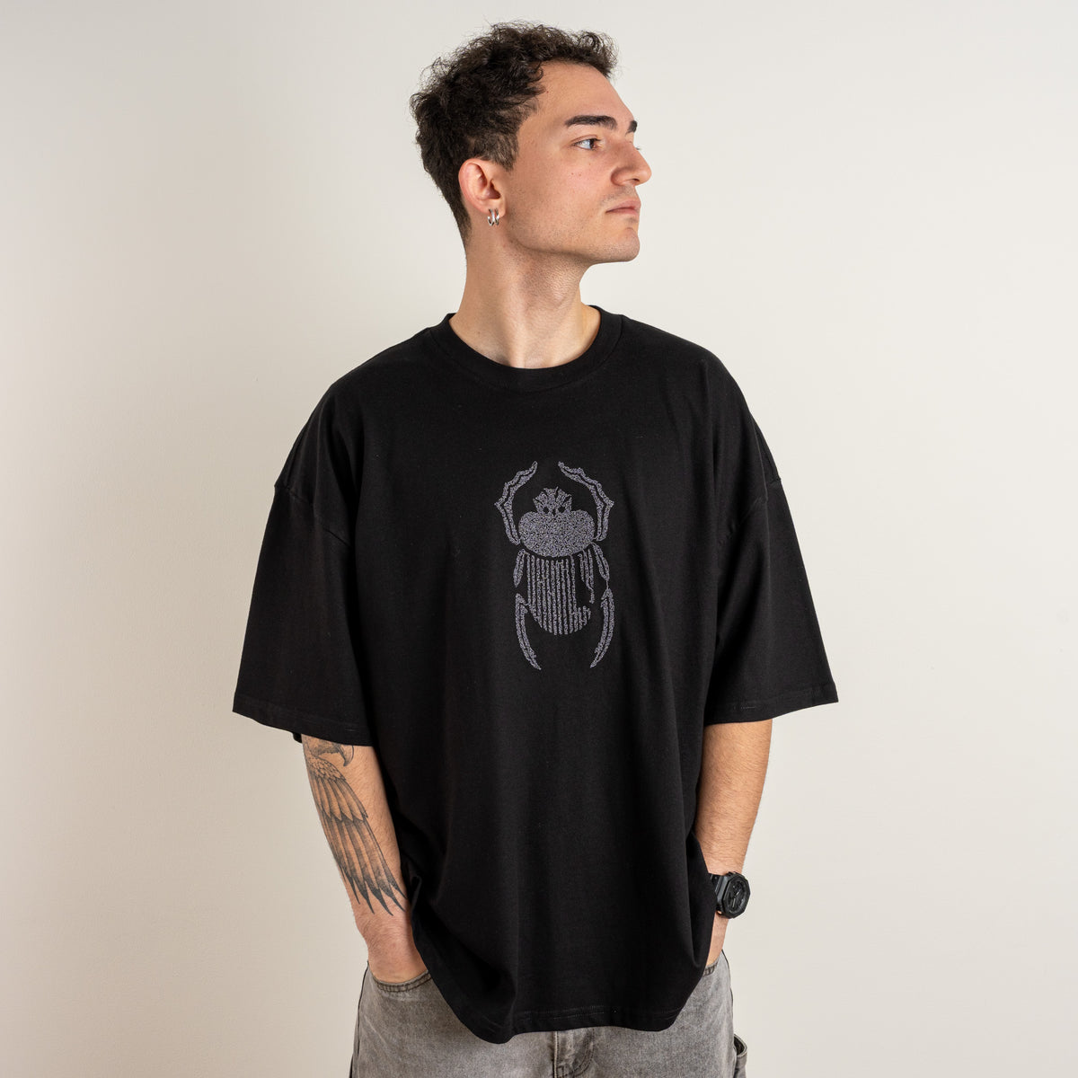 Three-quarter front view of a model wearing the Gordina The Scarab black oversized T-shirt with grey embroidery.