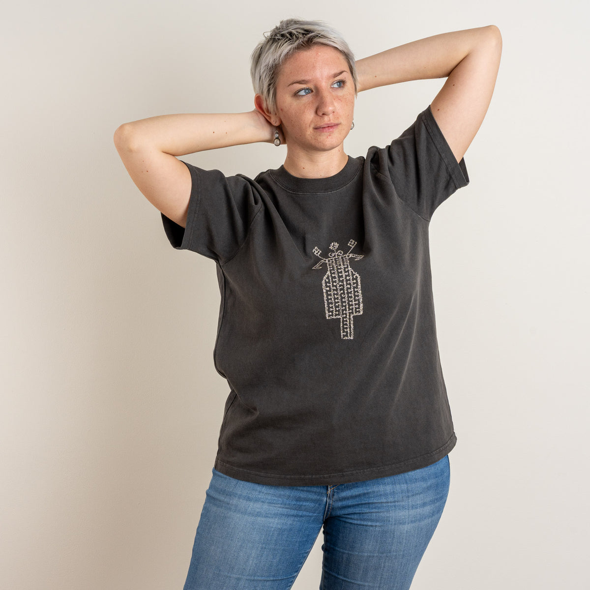 Woman with arms raised wearing The Ward Faded Black women's heavyweight cotton T-shirt with cream horse-and-rider embroidery, by Gordina, Netherlands.