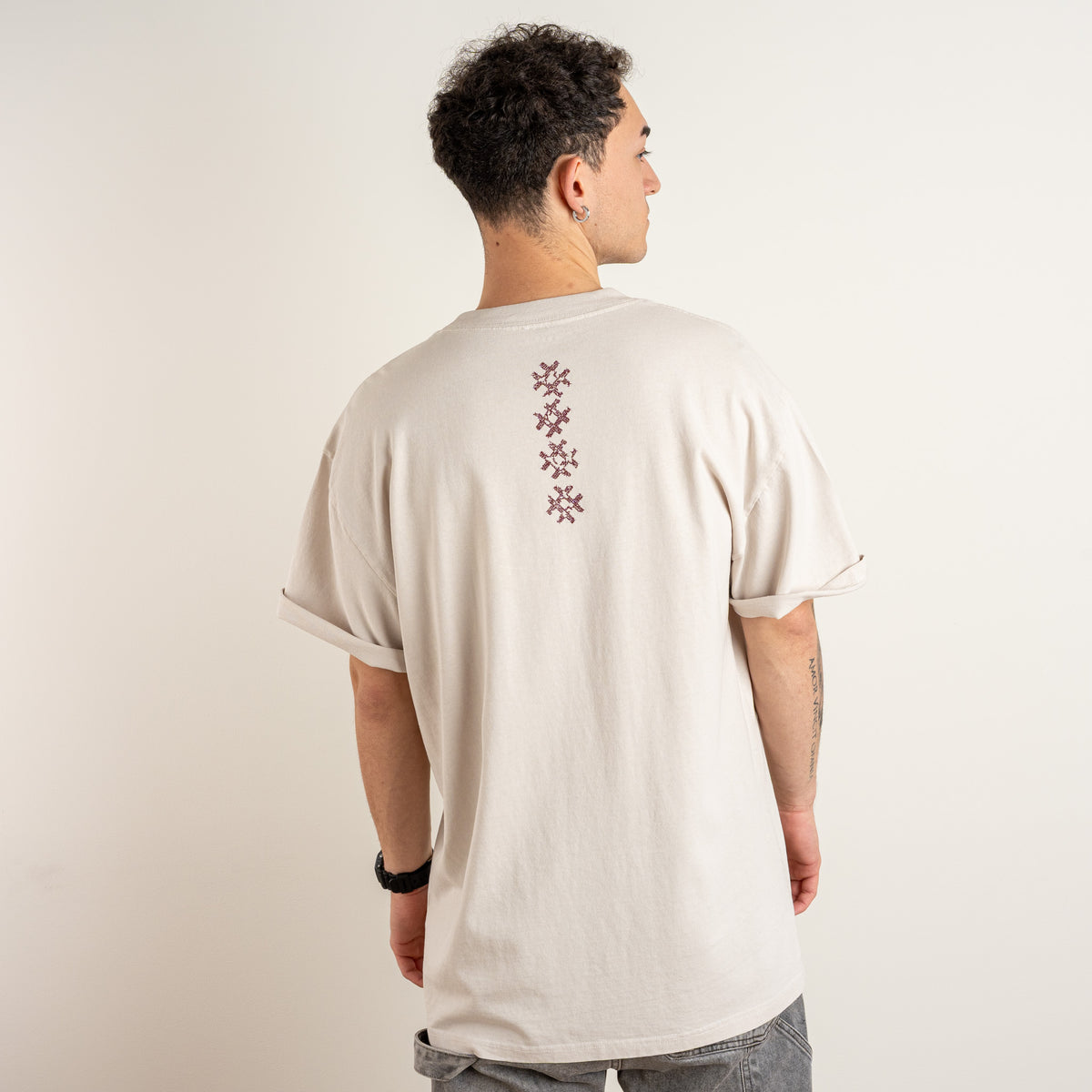 Back view of a model wearing the Gordina Unsigned Faded Beige T-shirt with vertical burgundy cross-stitch embroidery, made in the Netherlands.