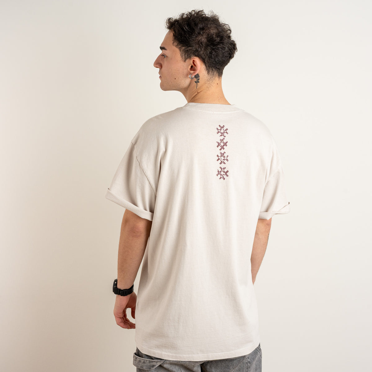 Side and back view of a model wearing the beige Gordina Unsigned T-shirt with burgundy cross embroidery, made in the Netherlands.