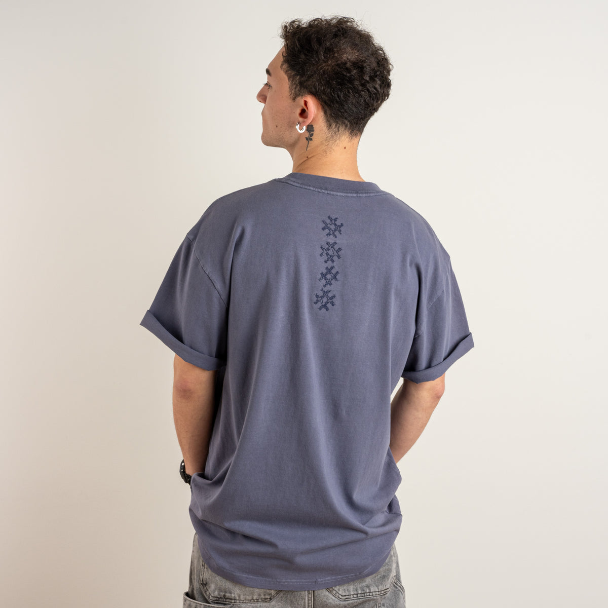 Back view of a model wearing the Gordina Unsigned Faded Indigo T-shirt with vertical navy cross embroidery, made in the Netherlands.