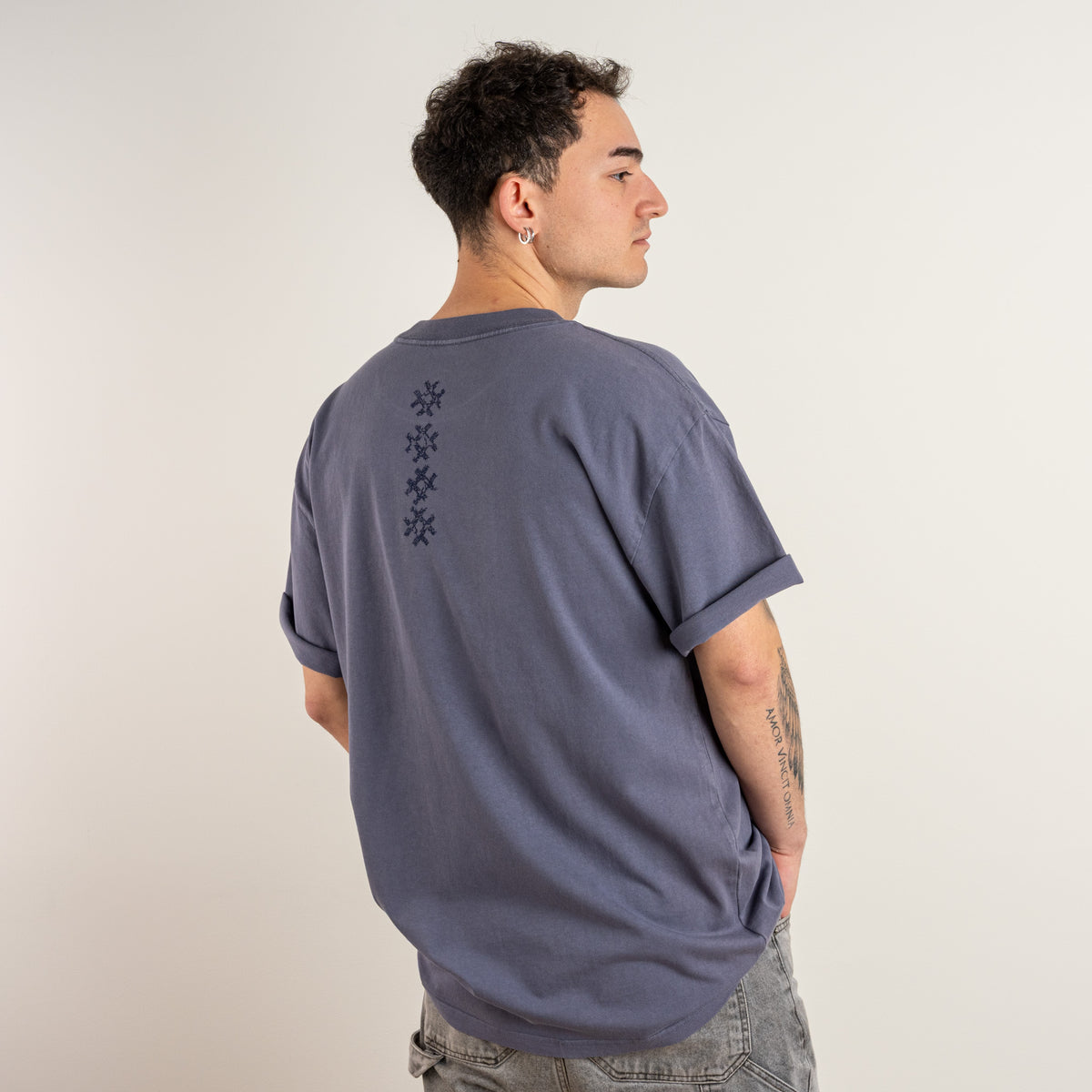 Three-quarter back view showing the navy embroidered cross motif on the Gordina Unsigned Faded Indigo T-shirt, embroidered in the Netherlands.