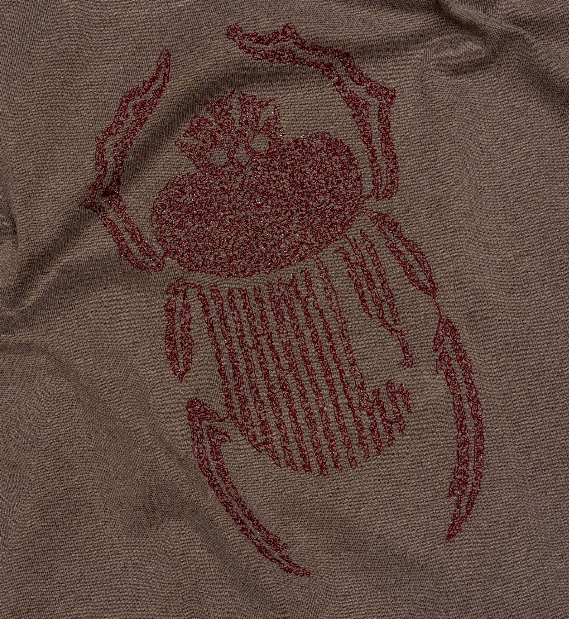 Angled detail of burgundy scarab embroidery stitched onto faded grey cotton.