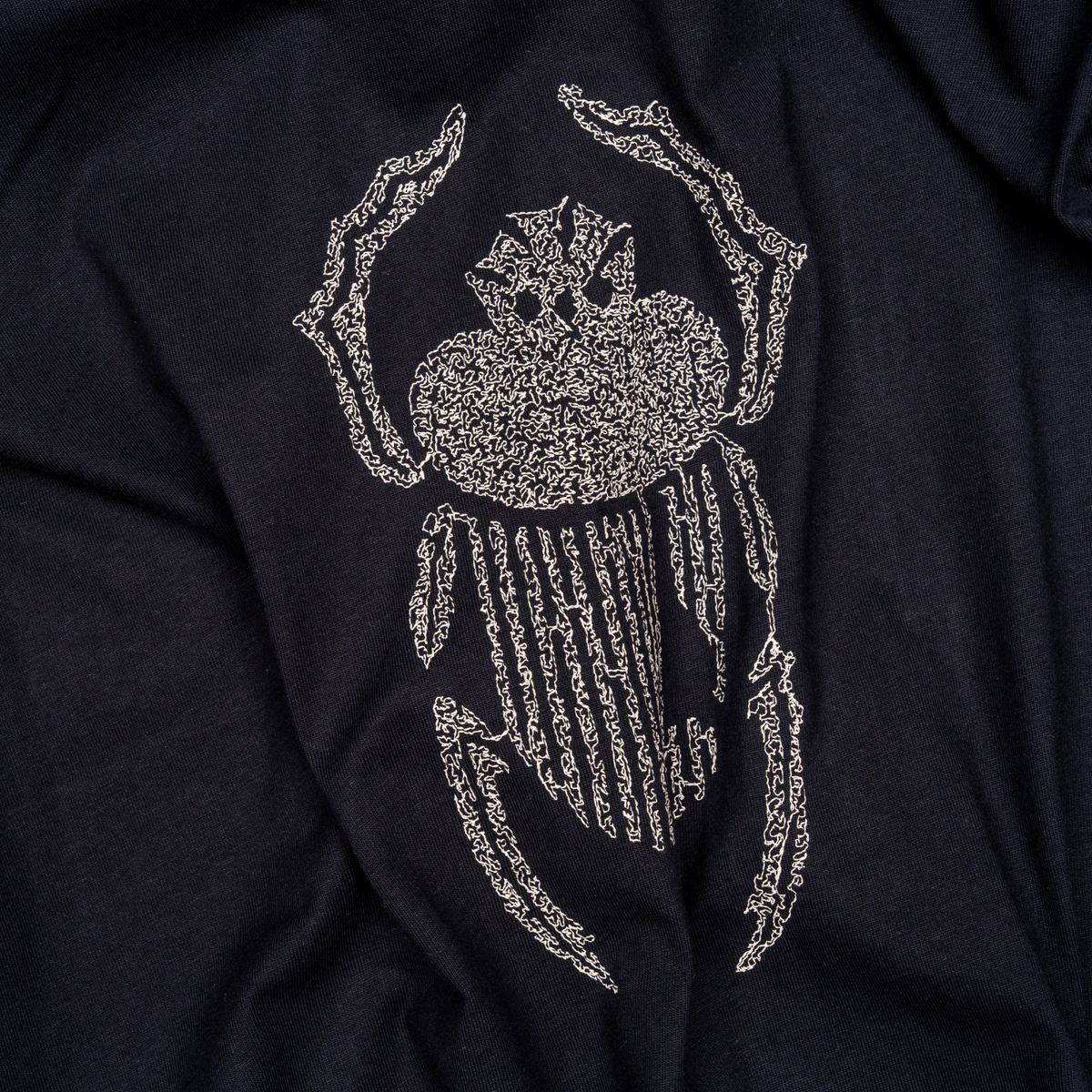 Angled macro detail of ivory scarab embroidery stitched onto navy cotton.