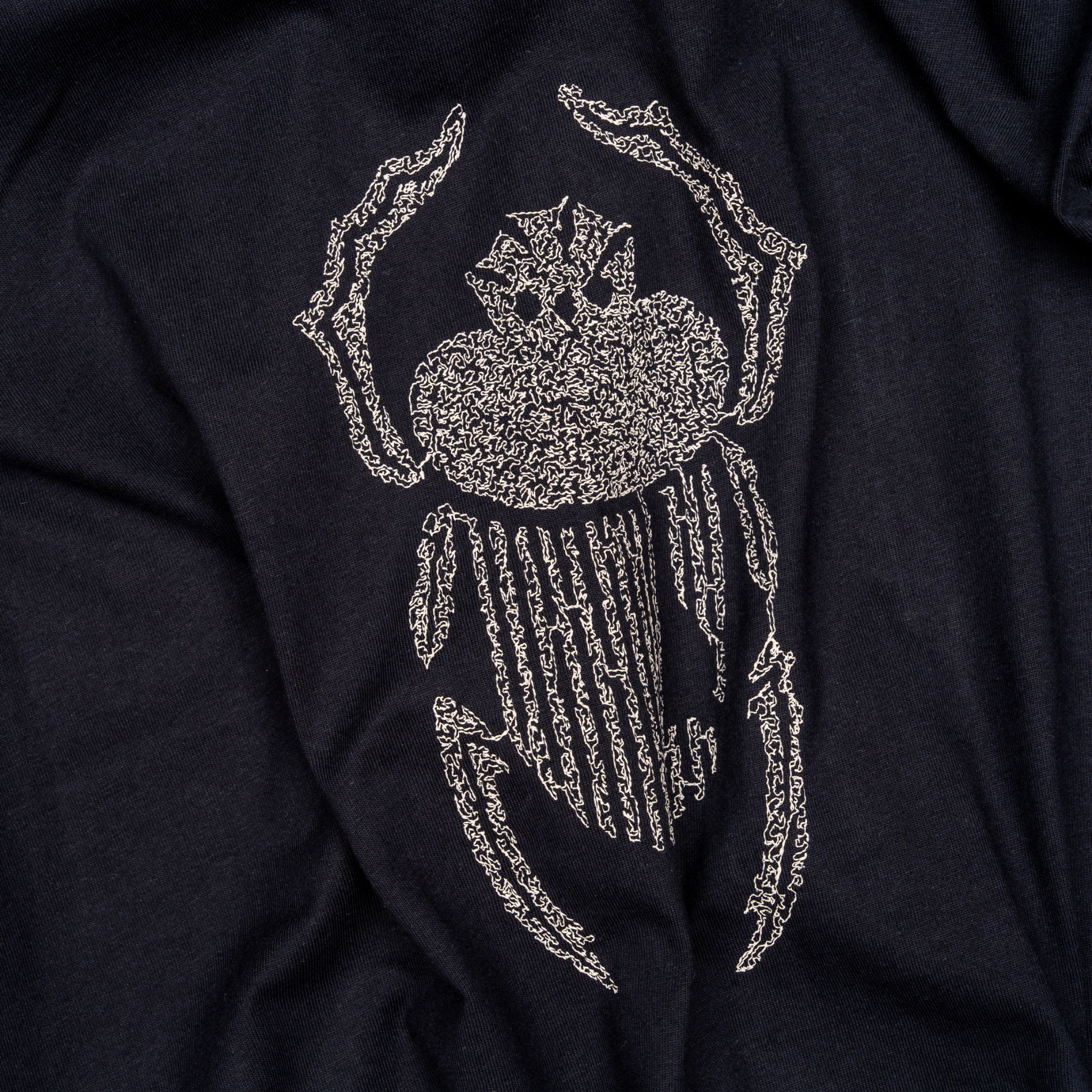 Angled macro detail of ivory scarab embroidery stitched onto navy cotton.