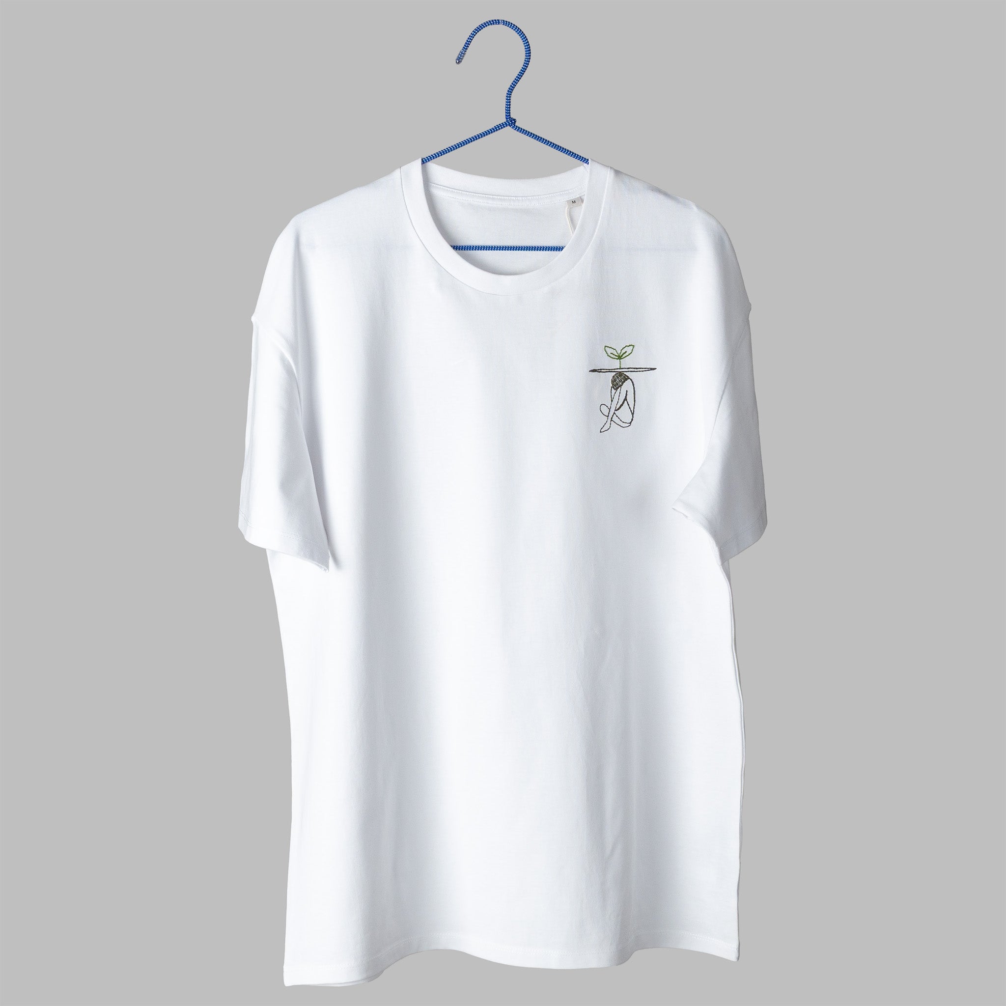A white t-shirt on a blue wire hanger against a light grey background. It features a small embroidered design on the left chest showing a line-art figure sitting huddled over, with a flat line above them and a small green plant sprouting from the top.