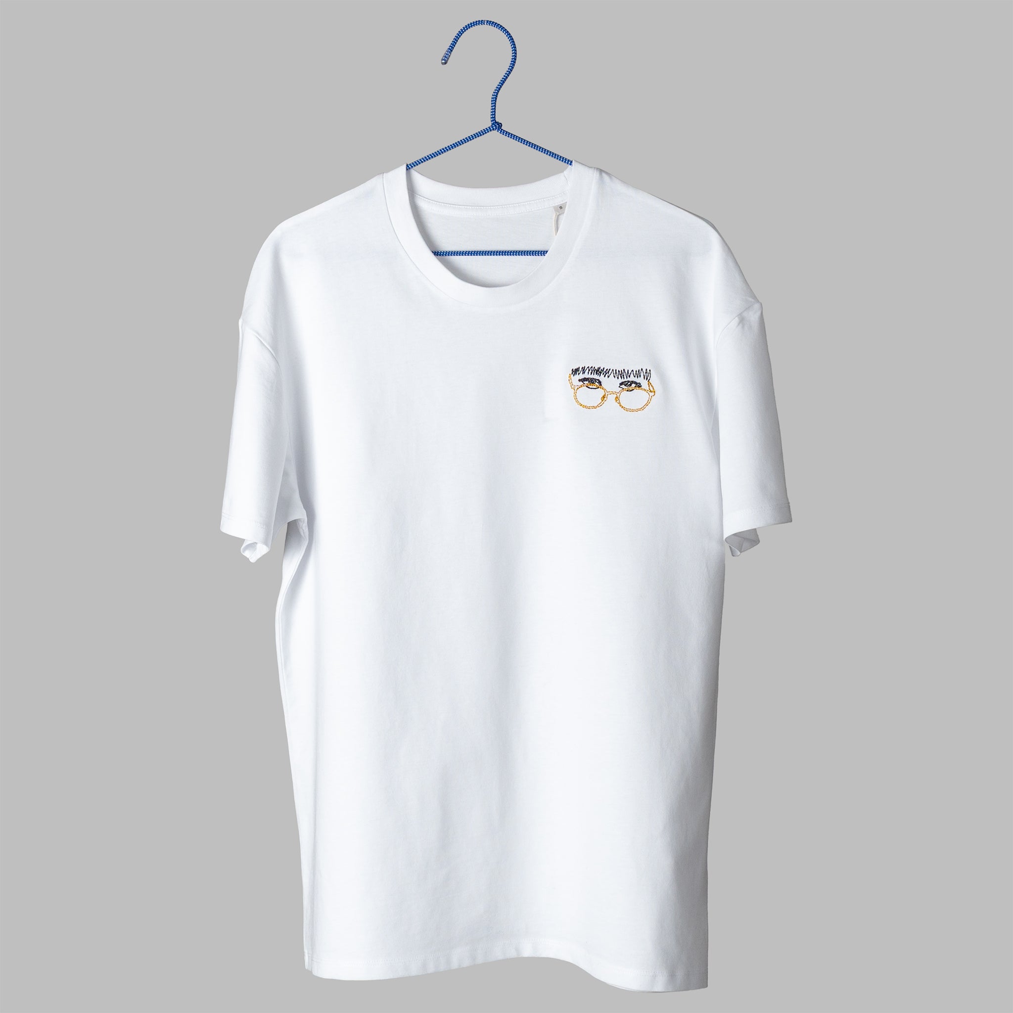A white t-shirt on a blue wire hanger against a light grey background. It features a small embroidered design on the left chest of a pair of eyes looking over yellow wire-rimmed glasses, topped with a thick, scribbled black unibrow.