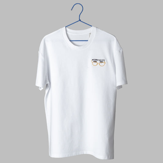 A white t-shirt on a blue wire hanger against a light grey background. It features a small embroidered design on the left chest of a pair of eyes looking over yellow wire-rimmed glasses, topped with a thick, scribbled black unibrow.