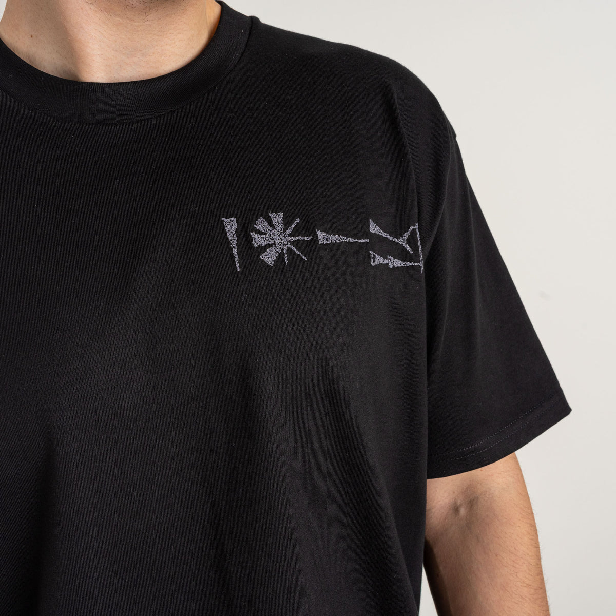 Detailed view of pale cuneiform-inspired embroidery on the chest of a black cotton T-shirt.