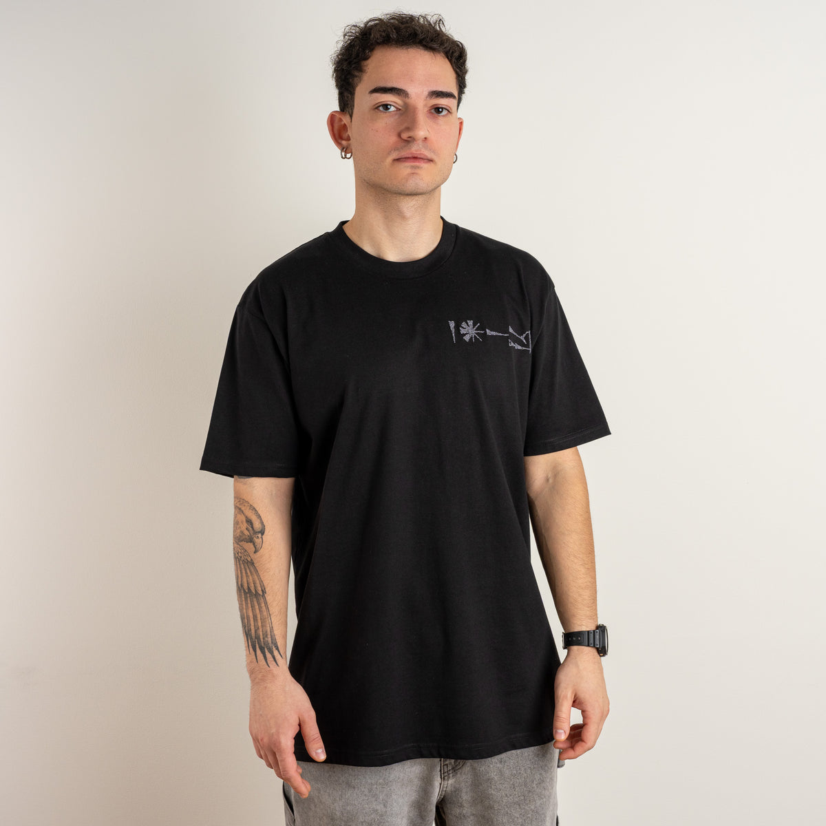 Model wearing the Gordina Undeciphered black T-shirt with a small cuneiform-inspired chest embroidery, front view.