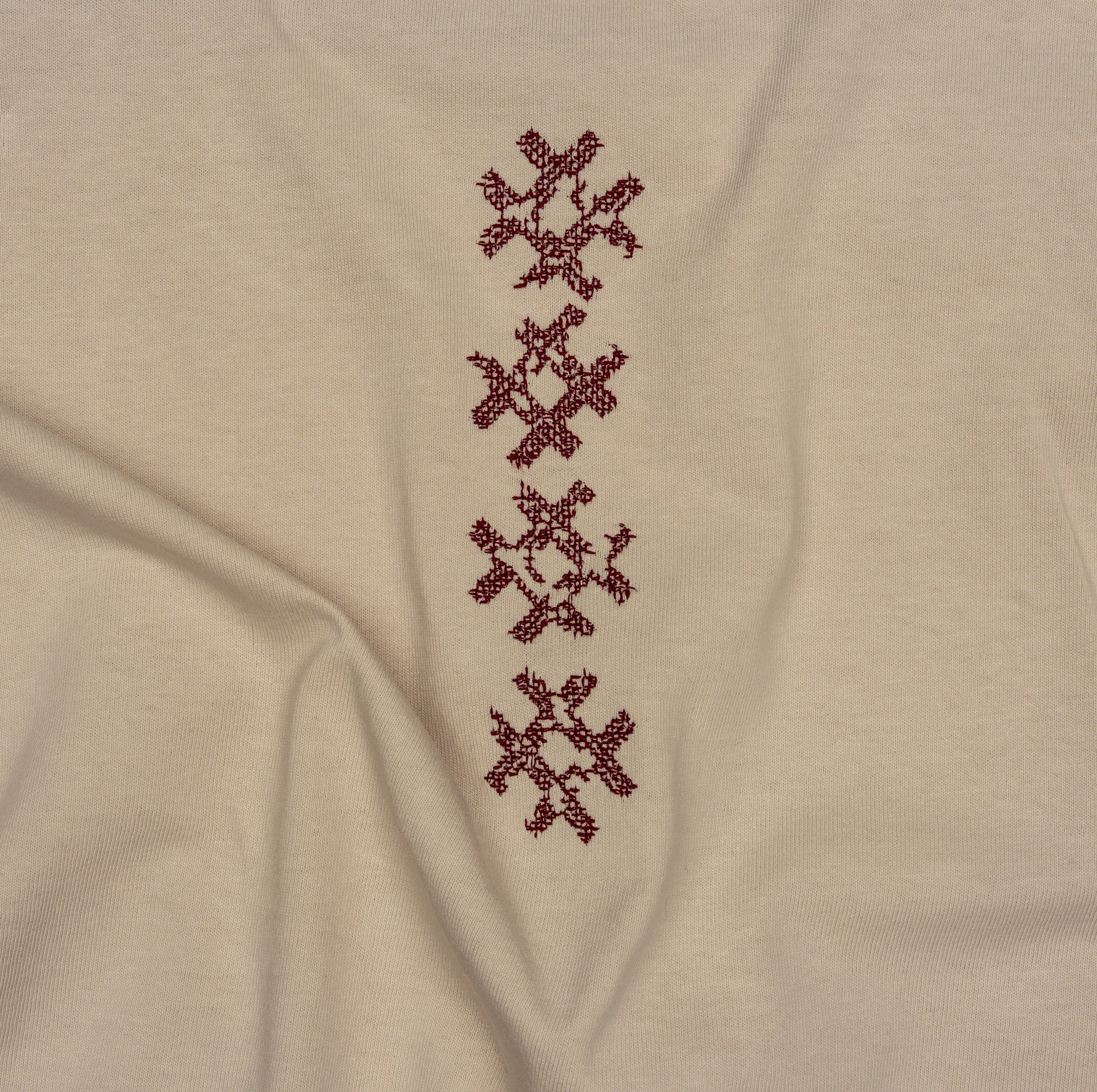 Angled close-up of burgundy cross-stitch embroidery on the beige Gordina Unsigned T-shirt.