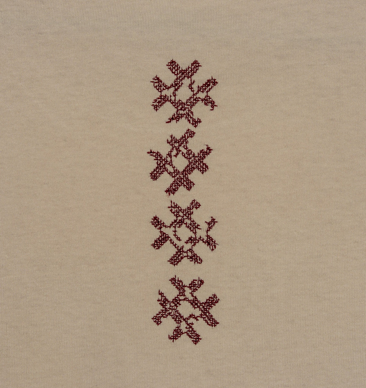 Close-up of the vertical burgundy cross-stitch motif on the beige Gordina Unsigned T-shirt.