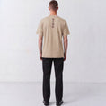 Load image into Gallery viewer, Back view of a model wearing the beige Gordina Unsigned T-shirt with vertical burgundy embroidery.
