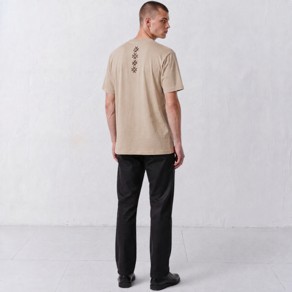 Three-quarter back view of a model wearing the beige Gordina Unsigned T-shirt with burgundy cross-stitch embroidery.