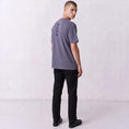 Load image into Gallery viewer, Three-quarter back view of a model wearing the Gordina Unsigned faded indigo T-shirt with navy cross embroidery.
