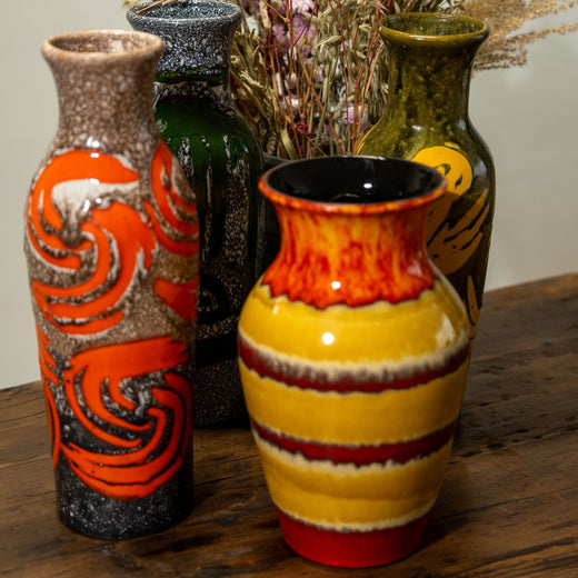 A collection of vintage ceramic vases displayed on a rustic wooden surface. Each vase features a unique glaze pattern with bold colors and textures, including vibrant yellows, deep greens, and fiery reds. The designs range from swirling abstract motifs to hand-painted organic elements. Dried flowers in the background add a natural, bohemian touch, enhancing the artisanal and nostalgic aesthetic of the arrangement.